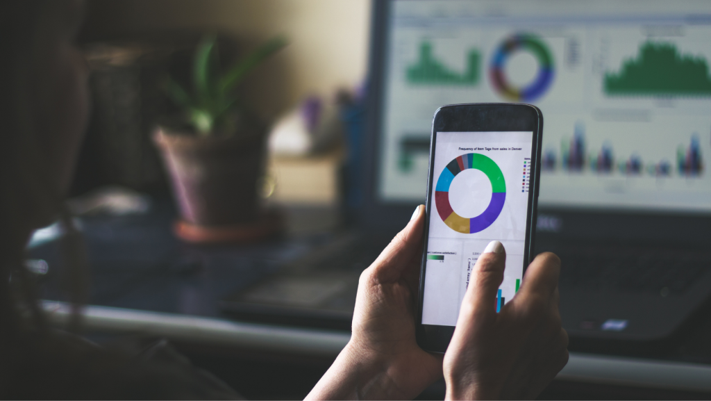 A person holding a smartphone displaying colorful data charts, with additional graphs visible on a laptop screen in the background.